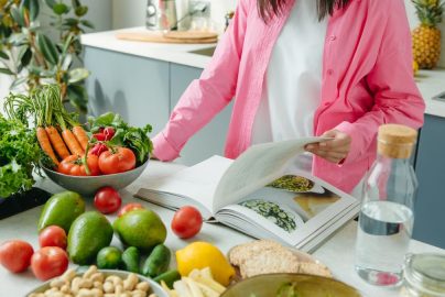 A person browsing a cookbook surrounded by fresh vegetables in a bright kitchen setting.