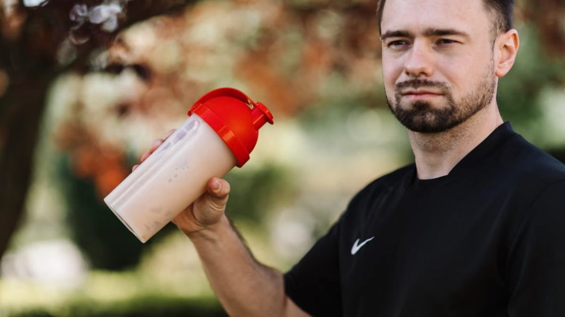 Adult man holds a protein shake in a red lid tumbler outdoors, wearing a black shirt.