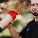 Adult man holds a protein shake in a red lid tumbler outdoors, wearing a black shirt.