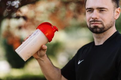 Adult man holds a protein shake in a red lid tumbler outdoors, wearing a black shirt.