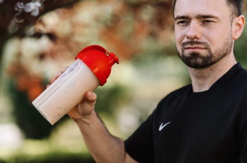 Adult man holds a protein shake in a red lid tumbler outdoors, wearing a black shirt.